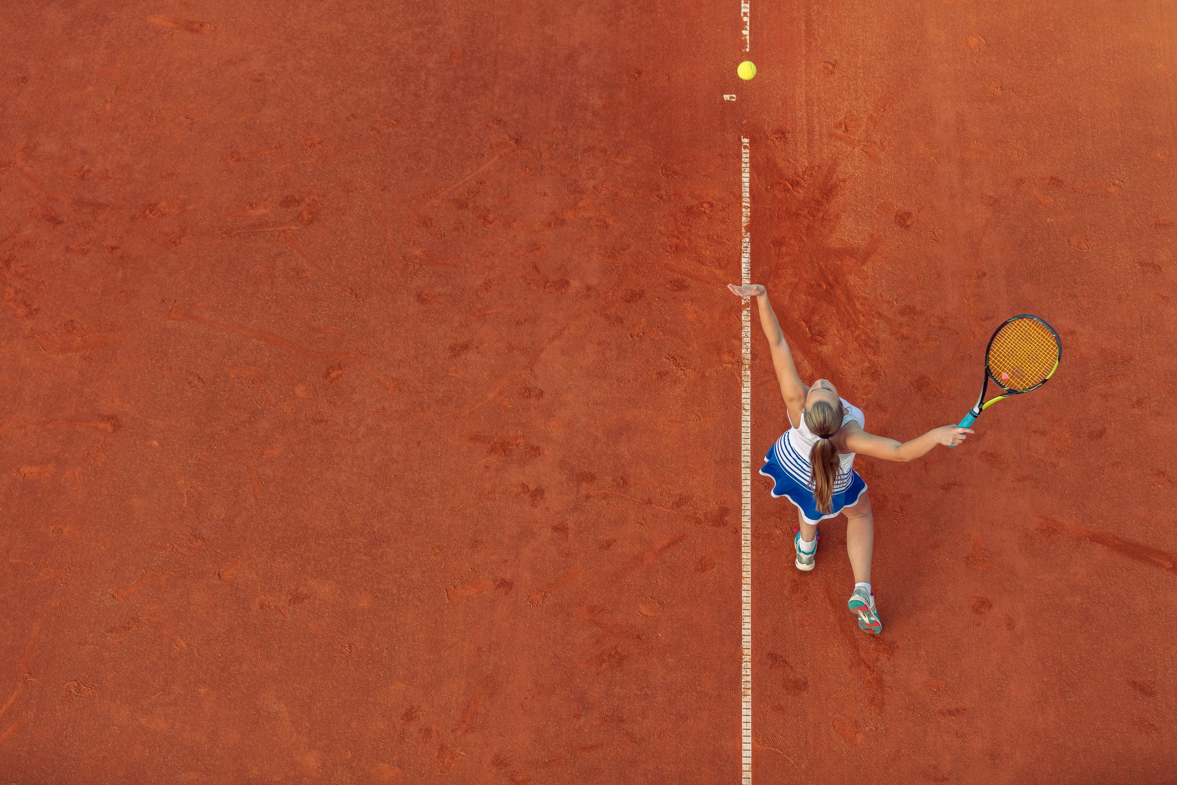 Aerial view of a woman in blue serving on a terracotta clay court