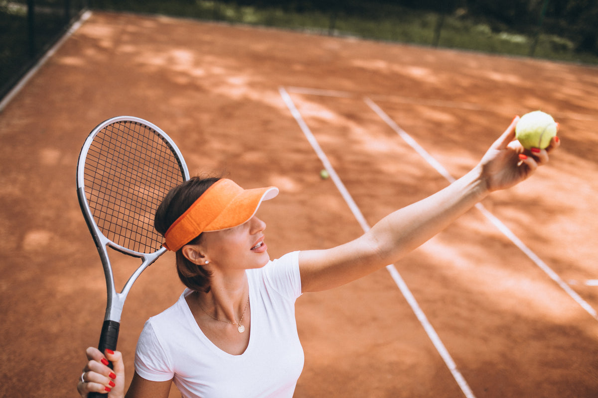 Woman in a white tee and orange visor tossing a ball to serve on a terracotta clay court