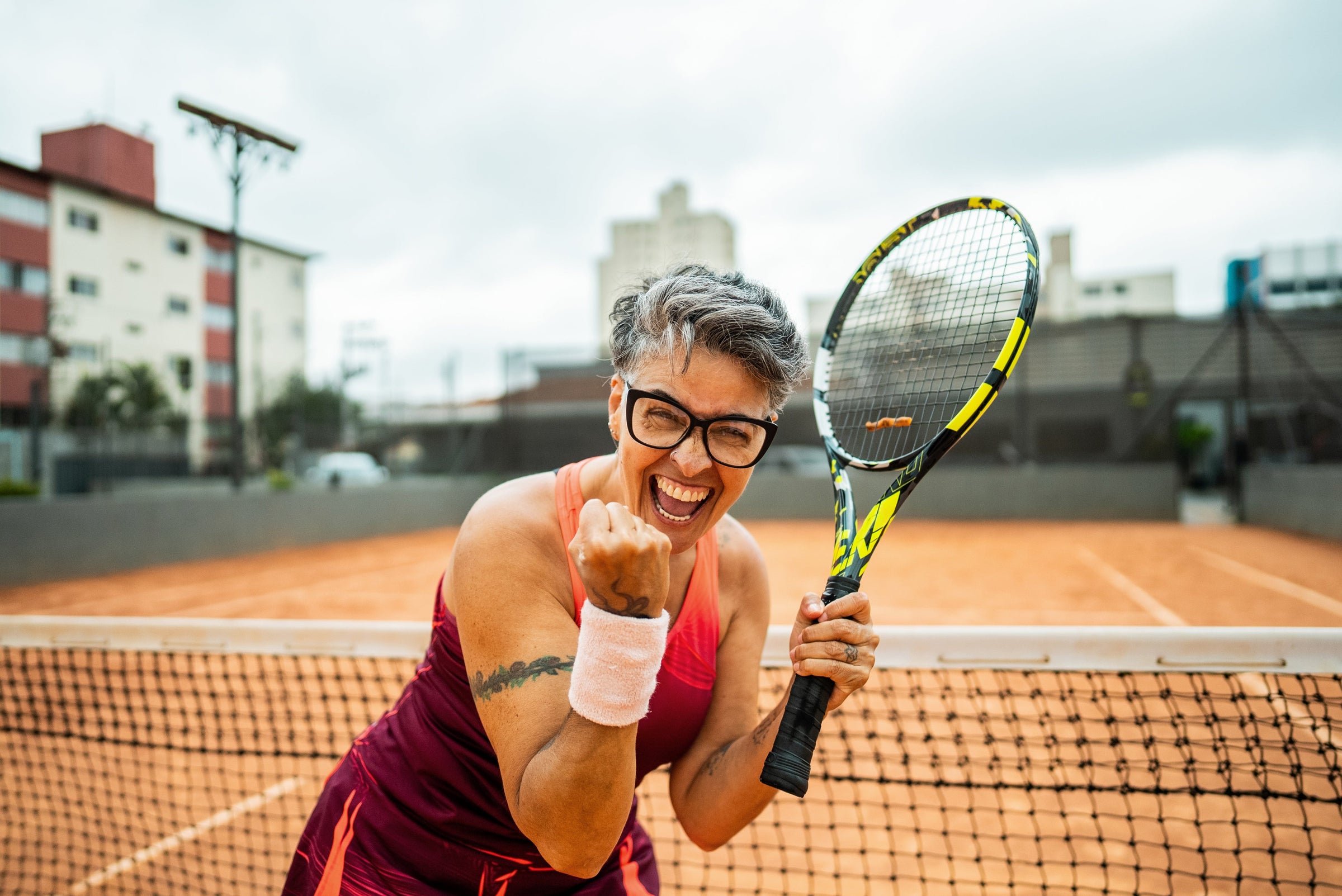 Smiling gray-haired woman in a coral tennis dress pumping her fist after a winning point at the net on a clay court