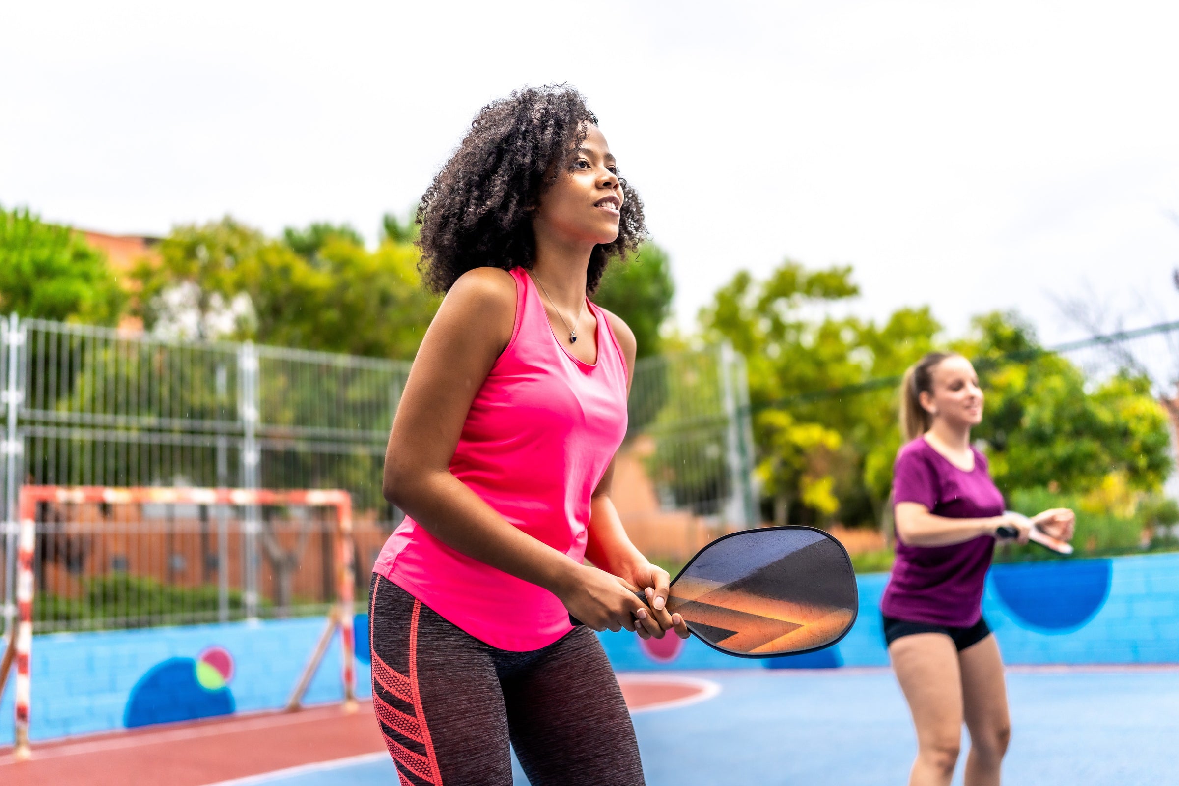 Two women in pink and purple playing pickleball doubles on a blue and orange court